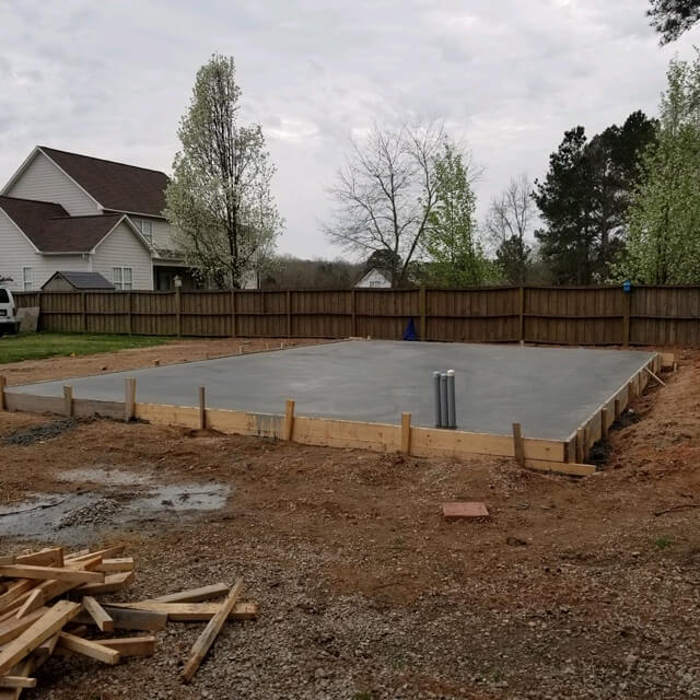 Freshly poured concrete foundation slab with wooden forms and utility pipes in a residential backyard